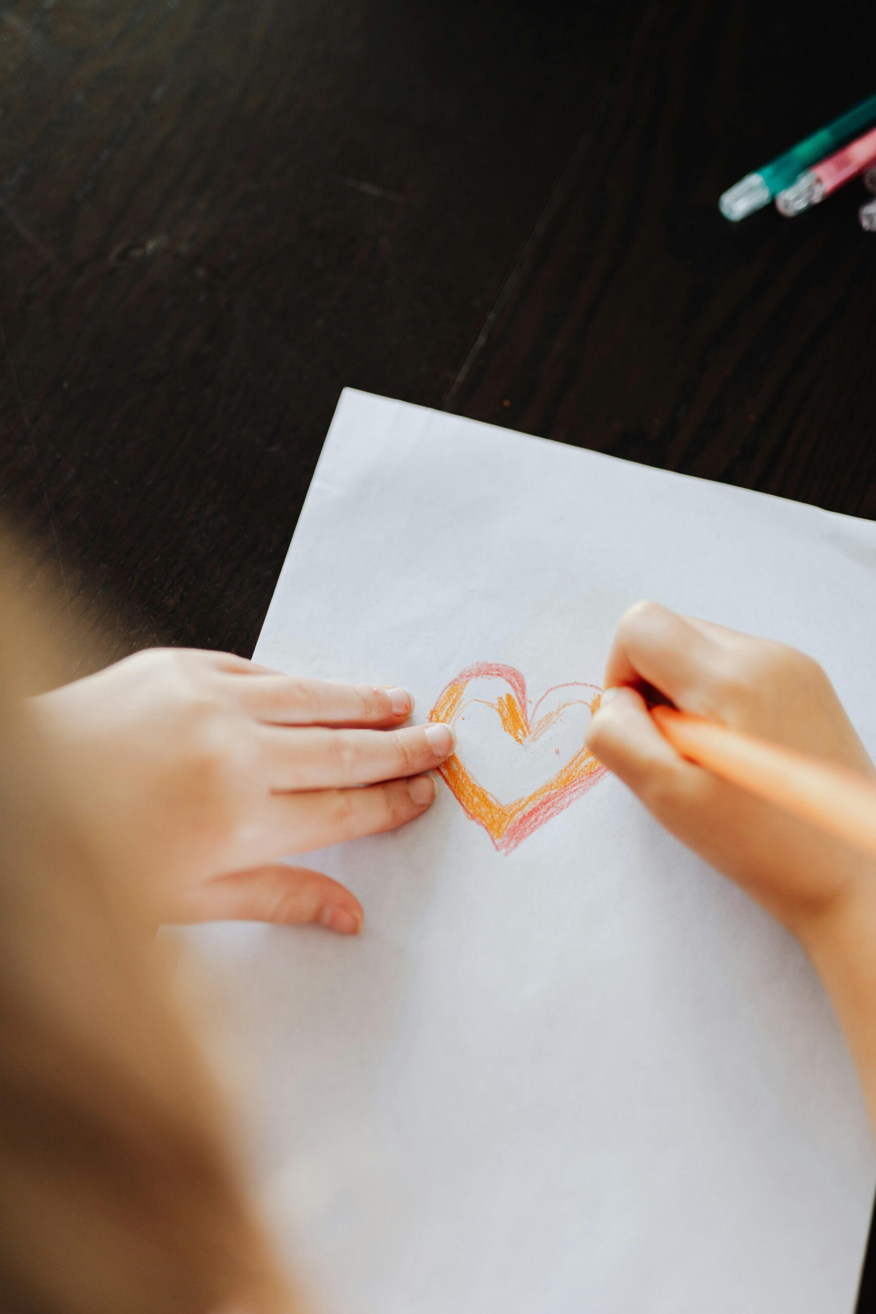 A child draws a colorful heart on white paper using colored pencils.