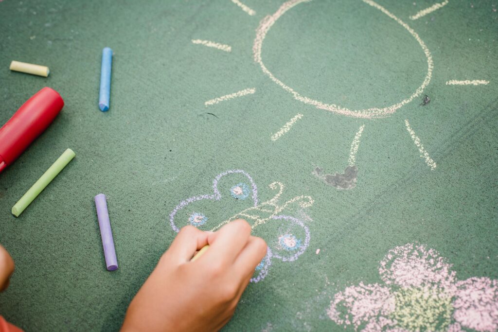 A child draws a sun and butterfly on pavement with colorful chalk sticks outdoors.